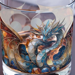 Op de afbeelding: Een helder glazen tumbler met een gedetailleerde illustratie van een draak. De draak heeft blauwe, gouden en oranje tinten. Het glas is ontworpen voor dranken en heeft een brede basis.