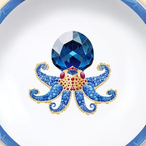 Peut inclure: Une assiette en céramique blanche avec une bordure bleue géométrique. L'assiette présente une illustration détaillée d'un poulpe bleu avec une grosse tête de gemme bleue à facettes, des accents rouges et des détails dorés.