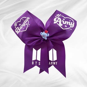 Pode incluir: Um laço de fita roxa com as palavras "Bangtan" e "Army" em branco. O laço apresenta um pequeno coala azul com uma flor vermelha. O texto "BTS" e "Army" também está impresso no laço.