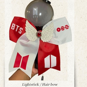 Può includere: Un lightstick con un fiocco rosso e bianco. Il fiocco presenta il testo "BTS" e vari loghi. Un ornamento bianco a forma di ala con una gemma rossa è centrato sul fiocco. Il lightstick è grigio con il logo "BTS".