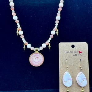 May include: A necklace and earrings set with a pink and white color scheme. The necklace features a pendant with a pink design and a beaded chain. The earrings are teardrop-shaped with a similar pink and white pattern. The tag reads "Handmade with love."