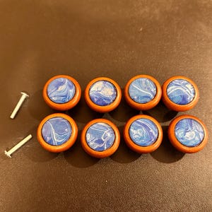 May include: Eight round cabinet knobs with a blue and white swirl design set in a wooden frame. Two silver screws are next to the knobs. The knobs are a decorative hardware item.