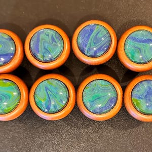 May include: Eight round wooden cabinet knobs with a marbled blue and green design. The knobs have a light brown wooden frame and a glossy, swirled center. The knobs are arranged in two rows of four.