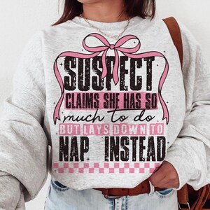 Może przedstawiać: Jasnoszara bluza z napisem "SUSPECT CLAIMS SHE HAS SO MUCH TO DO BUT LAYS DOWN TO NAP INSTEAD" w kolorze czarnym i różowym. Projekt zawiera różową wstążkę i wzór w szachownicę.