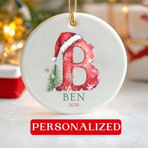 May include: A round white ceramic Christmas ornament featuring a large red letter B decorated with a Santa hat and a small green pine tree. The name BEN and the year 2026 are printed below the letter. The ornament hangs from a gold string.