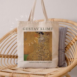 May include: A beige tote bag with a printed image of Gustav Klimt's "Portrait of Adele Bloch-Bauer I (1907)". The bag has black text that reads "Gustav Klimt" and "Portrait of Adele Bloch-Bauer I (1907)".