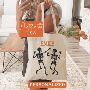 Puede incluir: Una bolsa de tela blanca con el nombre personalizado "Emily" y un gráfico de dos esqueletos bailando. La bolsa también tiene el texto "Printed in the USA" y "Personalized" impreso en ella.