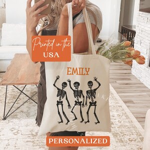 Puede incluir: Una bolsa de lona blanca con un nombre personalizado "Emily" y un gráfico de tres esqueletos bailando. La bolsa también dice "Impreso en los EE. UU." y "Personalizado".