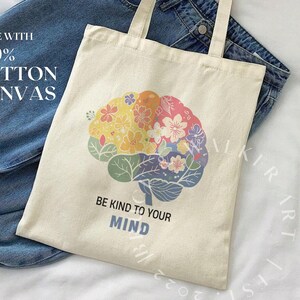May include: A beige tote bag made with 100% cotton canvas. The bag features a colorful brain design with flowers and the text "Be Kind to Your Mind".
