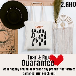 May include: A white tote bag with a black ghost design and the text "EMILY" and "2.GHOSTS". The bag is hanging on a wall with a macrame wall hanging and a black hat.