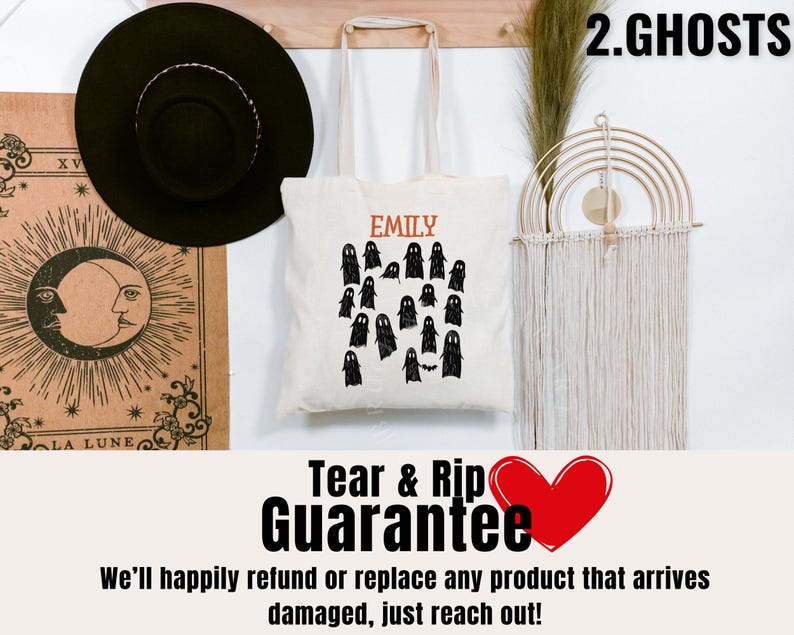 May include: A white tote bag with a black ghost design and the text "EMILY" and "2.GHOSTS". The bag is hanging on a wall with a macrame wall hanging and a black hat.