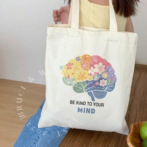 May include: A white tote bag with a colorful floral brain design and the text "Be kind to your mind".