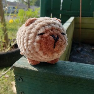 May include: A round, plush, crocheted animal figure made from soft, textured beige and light brown yarn. The creature features small rounded ears, a darker brown snout area, and a simple black stitched nose and mouth detail on its face.