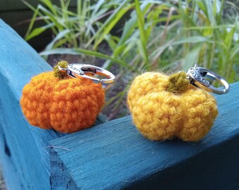 May include: Two crocheted pumpkin keychains, one orange and one yellow, sit on a blue surface. Each pumpkin has a green stem and a silver keychain ring. The pumpkins are detailed with textured yarn.