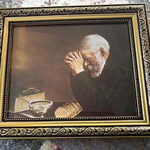 May include: Framed print featuring a man with a white beard and hair, hands clasped in prayer. The image includes a book, bowl, and bread. The frame is black and gold with ornate detailing.