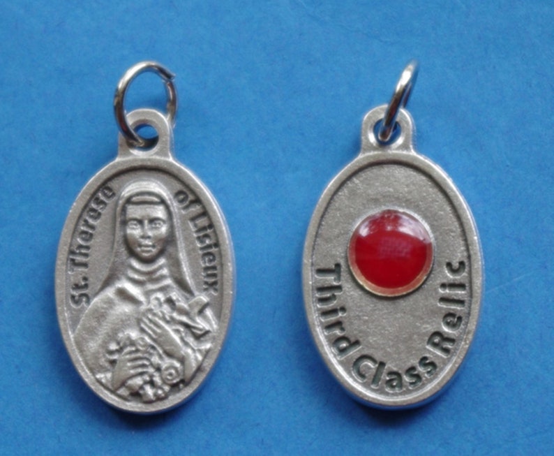 May include: Silver oval pendant with a picture of Saint Therese of Lisieux on one side and the words "Third Class Relic" and a red glass stone on the other side.