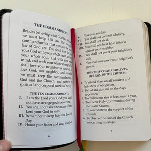 May include: Open book with black cover, displaying text about The Commandments and The Chief Commandments. The pages are white with black text, and a red ribbon bookmark is visible. The book is open to pages 6 and 7.