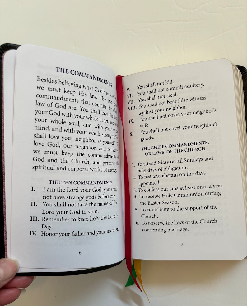 May include: Open book with black cover, displaying text about The Commandments and The Chief Commandments. The pages are white with black text, and a red ribbon bookmark is visible. The book is open to pages 6 and 7.