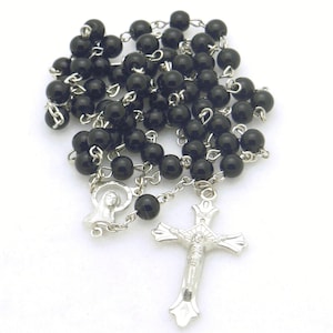 May include: A religious rosary featuring smooth, round black beads connected by silver-toned metal links. The piece includes a silver-toned crucifix pendant and a circular medal depicting a figure, all joined together in a continuous chain.