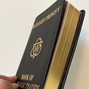 May include: Black leather-bound book titled "Blessed Trinity" and "Book of Catholic Prayers" with gold lettering and gilded page edges. The book has a gold emblem on the cover and colorful ribbon bookmarks.