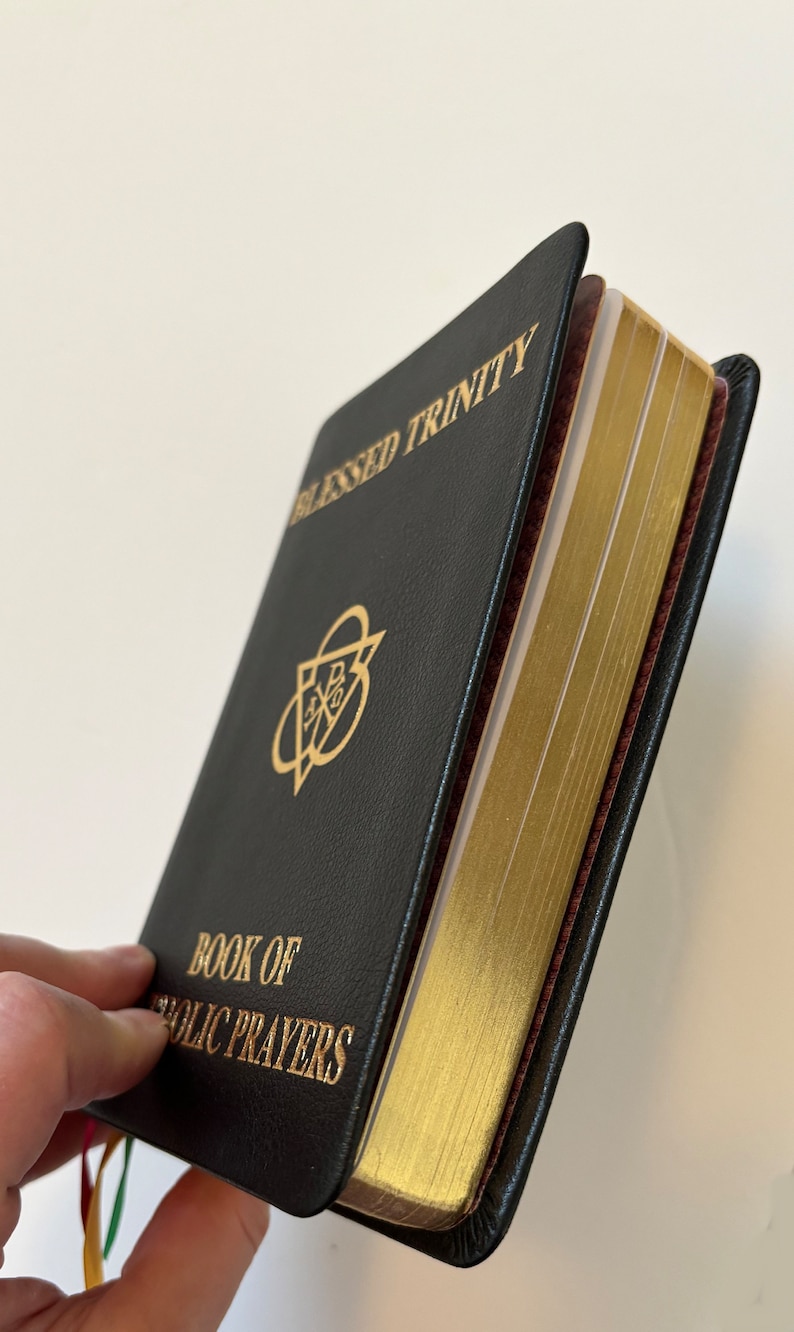 May include: Black leather-bound book titled "Blessed Trinity" and "Book of Catholic Prayers" with gold lettering and gilded page edges. The book has a gold emblem on the cover and colorful ribbon bookmarks.