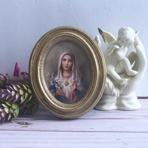 May include: An oval-shaped, gold-framed picture featuring a religious image of a woman with a halo, holding flowers and with a heart on her chest. A white ceramic angel figurine is to the right of the picture. Purple and green faux flowers are on the left.