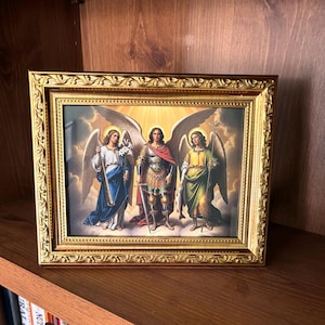 May include: A framed religious artwork featuring three figures with wings, set against a bright background. The figures are depicted in detailed attire, with one holding a sword and another a staff. The frame is ornate, with a gold finish.