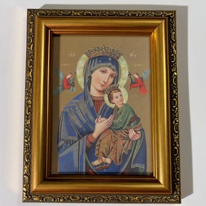 May include: A framed religious icon featuring a depiction of the Virgin Mary holding the Christ Child. The icon is in a gold-colored ornate frame. The Virgin Mary is wearing a blue robe and a crown. Two angels are on either side of the Virgin Mary.