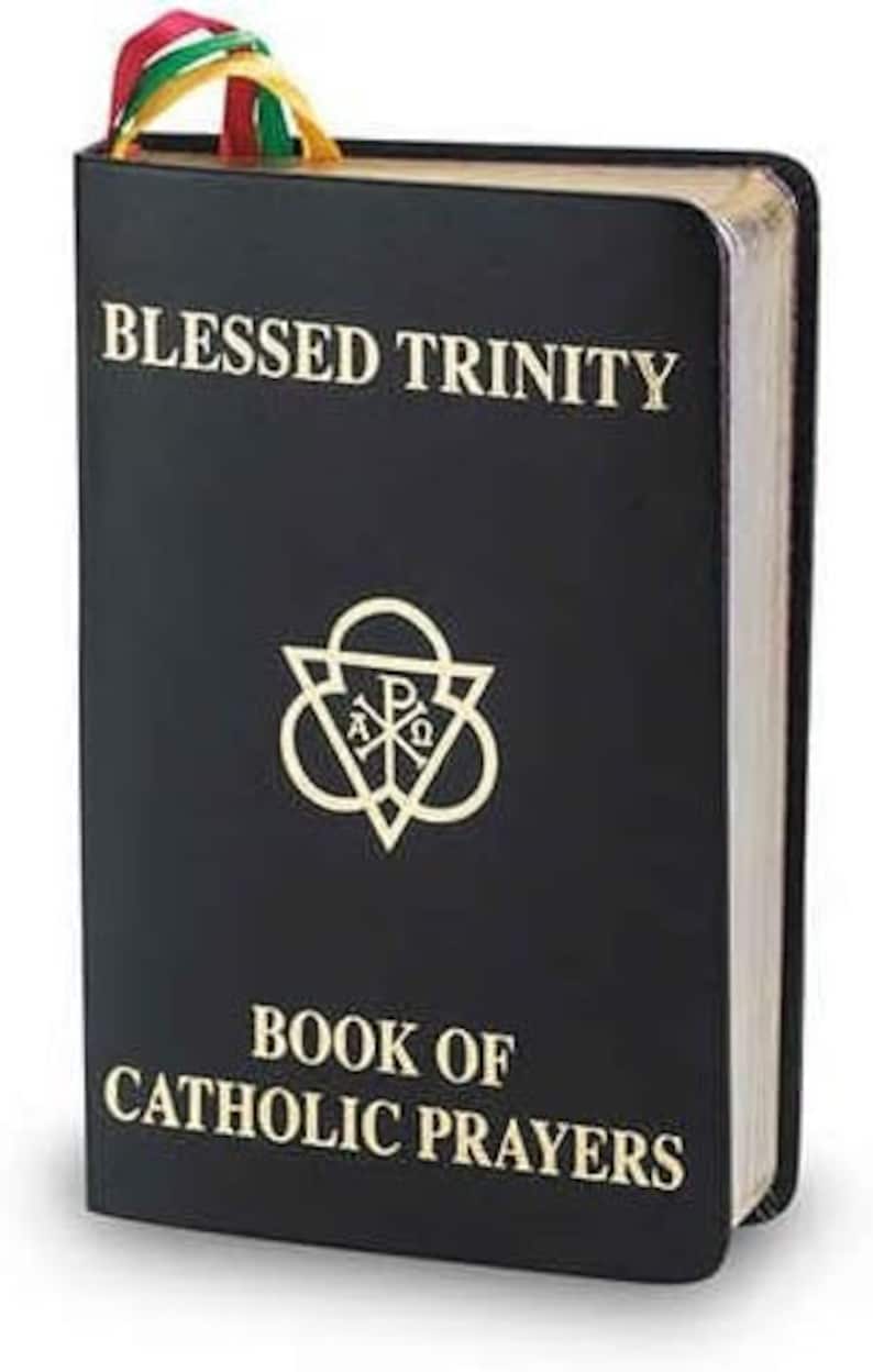 May include: A black book with gold lettering that reads "BLESSED TRINITY" and "BOOK OF CATHOLIC PRAYERS." A gold emblem is centered below the title. A colorful ribbon bookmark is visible at the top. The book's pages have a gold edge.