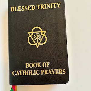 May include: Black leather-bound book titled "Blessed Trinity" and "Book of Catholic Prayers" in gold lettering. Features a gold emblem and a colorful ribbon bookmark. The book is closed and resting on a white surface.
