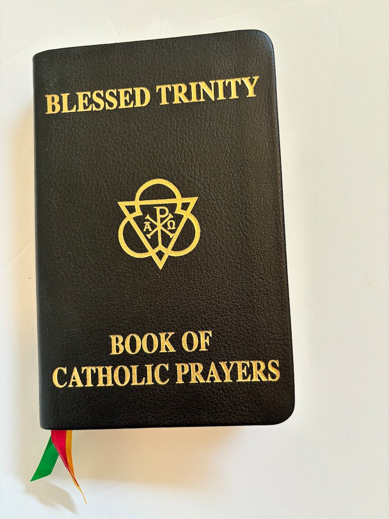 May include: Black leather-bound book titled "Blessed Trinity" and "Book of Catholic Prayers" in gold lettering. Features a gold emblem and a colorful ribbon bookmark. The book is closed and resting on a white surface.
