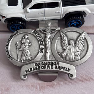 May include: Silver car charm with a religious theme. It features a depiction of Saint Christopher, Jesus on the cross, and an angel. The charm also includes the text "Protect Us" and "Grandson Please Drive Safely".