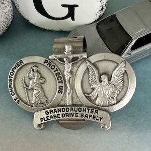 May include: Silver car charm with a silver cross, a silver angel, and a silver image of St. Christopher. The charm has a silver banner that reads "Granddaughter Please Drive Safely" and "Protect Us".