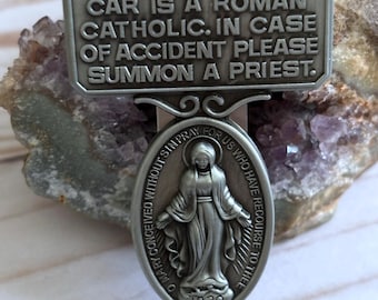 May include: A silver car charm with a religious message: "The owner of this car is a Roman Catholic. In case of accident please summon a priest." The charm also features a silver oval with an image of the Virgin Mary and the text "O Mary conceived without sin pray for us who have recourse to thee 1830."