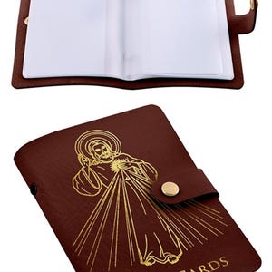 May include: A brown faux leather holder for holy cards featuring a gold foil embossed image of Divine Mercy and the text HOLY CARDS on the front cover. The item includes a gold snap button closure and internal clear plastic sleeves for storage.