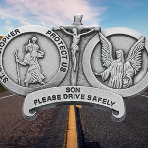 May include: A silver-colored car medallion with religious imagery. It features St. Christopher carrying a child, a crucifix, and an angel. The text reads "PROTECT US SON PLEASE DRIVE SAFELY" and "ST. CHRISTOPHER". The background shows a road and mountains.