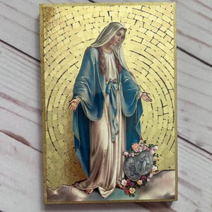 May include: A rectangular religious icon featuring a depiction of a woman in a blue robe and white veil, with a gold mosaic background. The image includes two medallions and flowers at the bottom. The overall color scheme is gold, blue, and white.