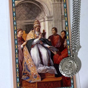 May include: A rectangular laminated prayer card featuring a religious painting of a pope in ornate robes surrounded by figures, paired with a round silver-toned metal pendant on a silver chain, inscribed with Saint Gregory Pray For Us.