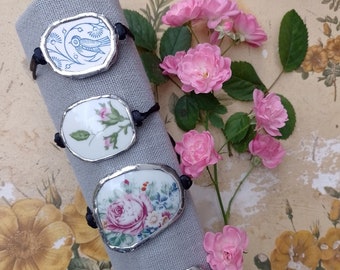May include: Four bracelets with black cords and silver-rimmed porcelain charms featuring floral and bird designs. The charms are displayed on a gray fabric background with pink roses.