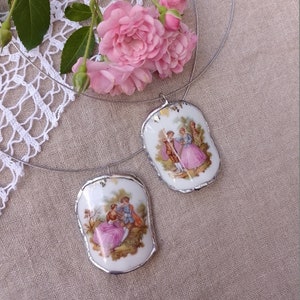 May include: A silver necklace with two porcelain pendants featuring vintage illustrations of couples in a garden setting. The pendants have gold accents and are attached to a thin silver chain.