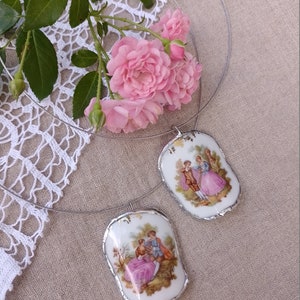 May include: A silver necklace with two porcelain pendants featuring vintage illustrations of couples in a garden setting. The pendants have a slightly irregular, hand-painted look.