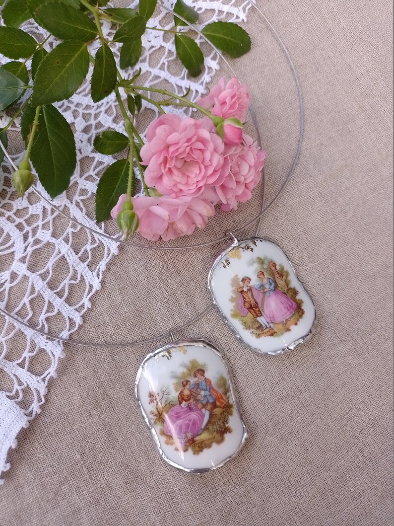 May include: A silver necklace with two porcelain pendants featuring vintage illustrations of couples in a garden setting. The pendants have a slightly irregular, hand-painted look.