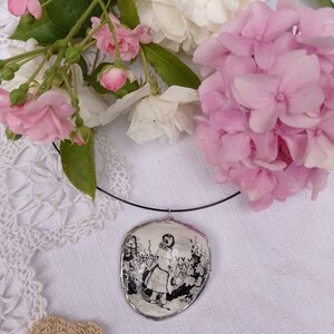 Pu&ograve; includere: Una collana con un cammeo in bianco e nero che raffigura una ragazza con un vestito che tiene un annaffiatoio. La collana &egrave; su uno sfondo bianco con fiori rosa.