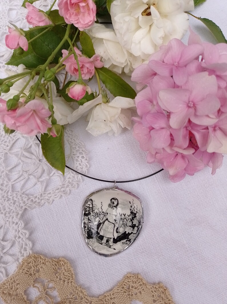 Pu&ograve; includere: Una collana con un cammeo in bianco e nero che raffigura una ragazza con un vestito che tiene un annaffiatoio. La collana &egrave; su uno sfondo bianco con fiori rosa.