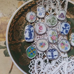 May include: A collection of 12 vintage porcelain charms, each with a different floral or geometric design. The charms are set in silver-toned metal frames and are displayed on a green and gold plate with a white lace doily.