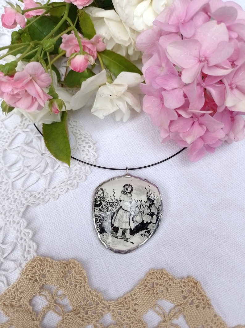 Pu&ograve; includere: Una collana con un ciondolo d'argento che raffigura un'illustrazione in bianco e nero di Alice nel Paese delle Meraviglie. Il ciondolo &egrave; appeso a un cordoncino nero ed &egrave; circondato da pizzo bianco e fiori rosa.