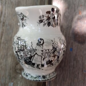 Pu&ograve; includere: Un'anfora di ceramica bianca con un motivo floreale bianco e nero e una scena di bambini che giocano. L'anfora ha un manico e un beccuccio.