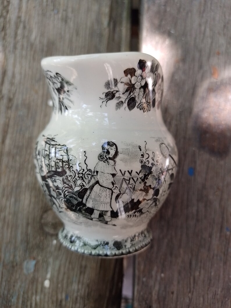 Pu&ograve; includere: Un'anfora di ceramica bianca con un motivo floreale bianco e nero e una scena di bambini che giocano. L'anfora ha un manico e un beccuccio.
