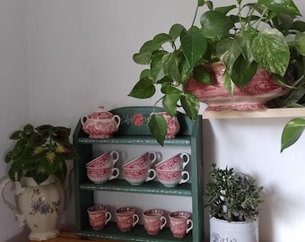 May include: A green wooden shelf unit displays six teacups and a sugar bowl, all with a red and white floral pattern. A large potted plant sits on top, and other plants are in decorative pots.