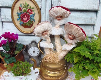 May include: Three whimsical mushroom-shaped figures with painted faces and white lace trim, arranged in a vintage brass teapot with straw and a fern plant in the background.